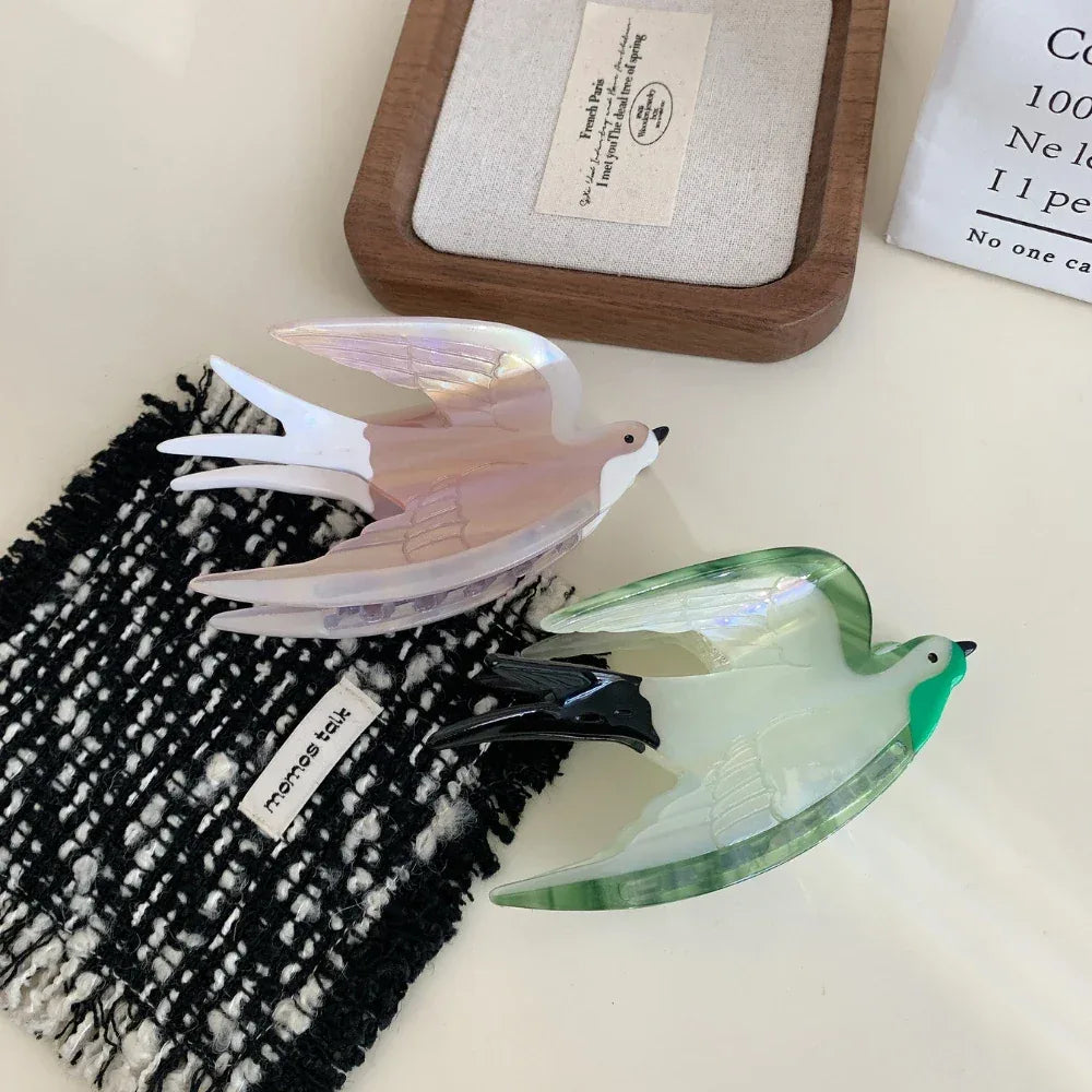 Two acrylic bird hair clips, one pearlescent pink and white, the other green and white, displayed on a textured black and white cloth.