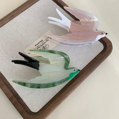 Two acrylic bird hair clips, one pearlescent pink and white, the other green and white, placed on a wooden tray with a linen insert.
