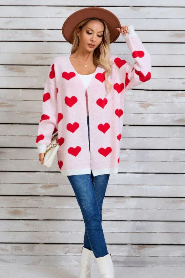 Woman wearing a pink longline cardigan with a red heart print, a white tank top, blue jeans, and a brown hat.