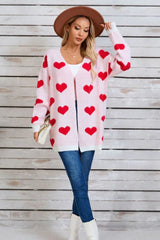 Woman wearing a pink longline cardigan with a red heart print, a white tank top, blue jeans, and a brown hat.