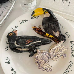 A close-up of three decorative bird hair clips on a plate, showcasing a toucan, a crow, and a peacock design.