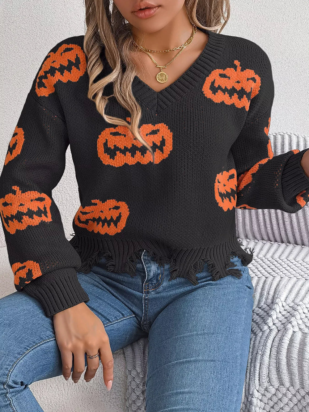 Close-up of a woman wearing a black long-sleeve v-neck cropped knit sweater with an all-over orange pumpkin print and a distressed hem, sitting on a couch.
