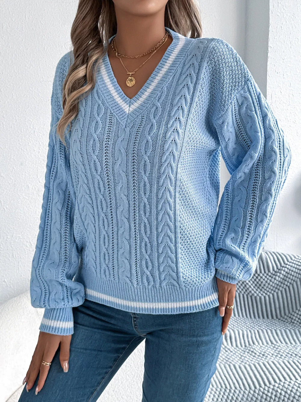 front view of a woman wearing a light blue cable knit v-neck sweater with white stripes, blue jeans, and gold necklaces