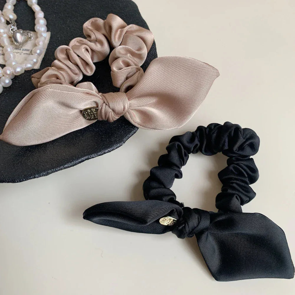 Beige and black satin scrunchies with bow details and gold-tone charms on a black textured surface