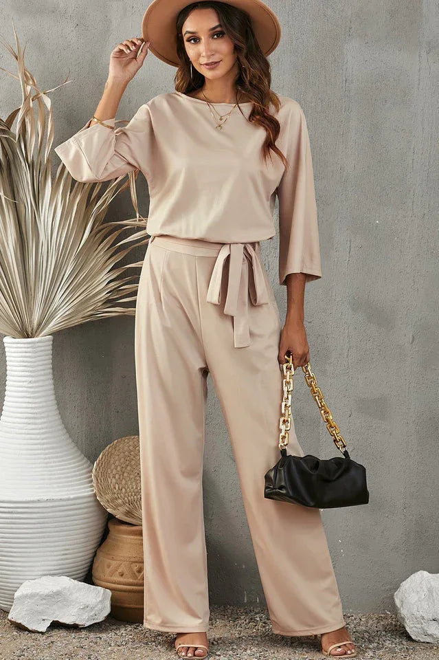 woman wearing a beige wide leg jumpsuit with a tie waist and holding a black clutch