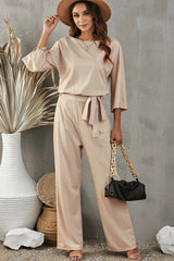woman wearing a beige wide leg jumpsuit with a tie waist and holding a black clutch
