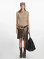 Woman wearing a beige long-sleeve knit top, an olive green faux leather skirt with an asymmetrical hem, a beige beanie, and brown boots, holding a black tote bag.