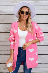 Woman wearing a pink longline cardigan with a white heart print, a white tank top, and blue jeans, holding a coffee cup.