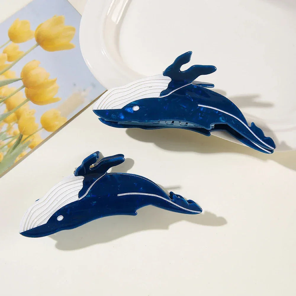 Two blue and white whale-shaped hair claw clips placed on a white surface with a floral picture in the background.