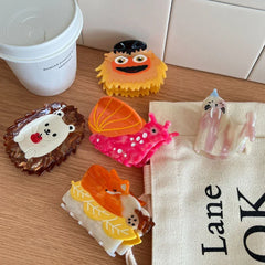 A group of decorative hair clips including a fox, hedgehog, snail, seal, and a fuzzy character, placed near a coffee cup and a canvas bag.