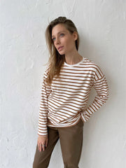 Woman wearing a white and brown striped long-sleeve top and brown faux leather pants, posing against a textured white wall.