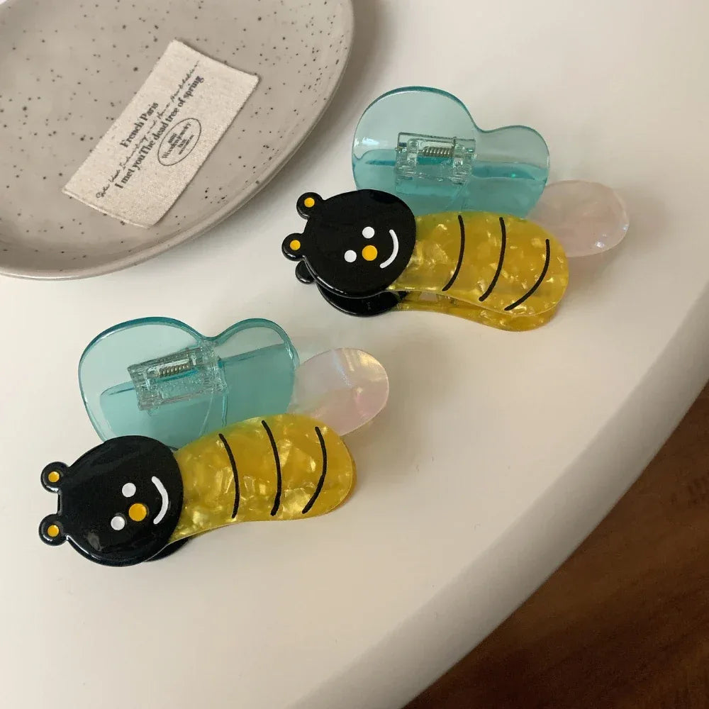 Two bee-shaped hair clips with translucent blue wings and yellow striped bodies arranged on a white surface.