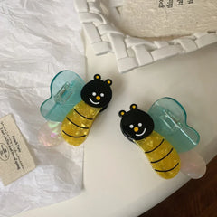 Two bee-shaped hair clips with translucent blue wings, scattered on a white textured surface.