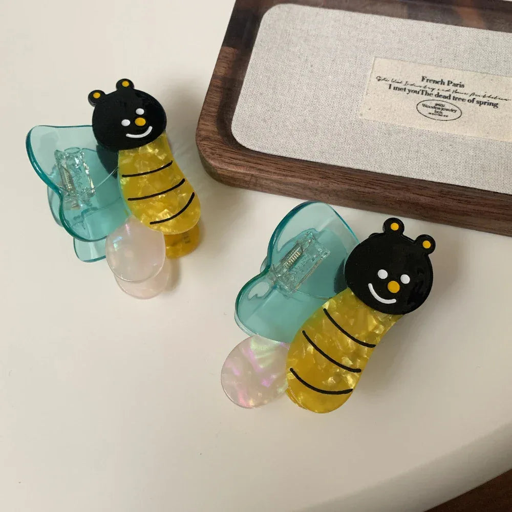 Two bee-shaped hair clips with blue wings, resting on a white surface next to a wooden tray.