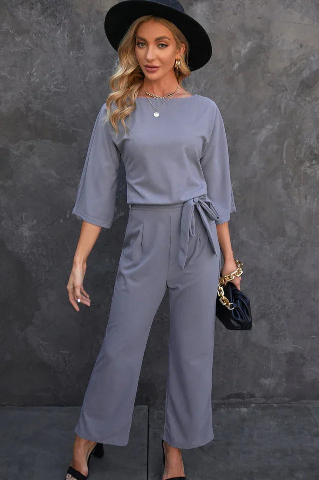 woman wearing a grey wide leg jumpsuit with a tie waist and a black hat