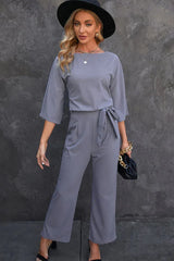 woman wearing a grey wide leg jumpsuit with a tie waist and a black hat