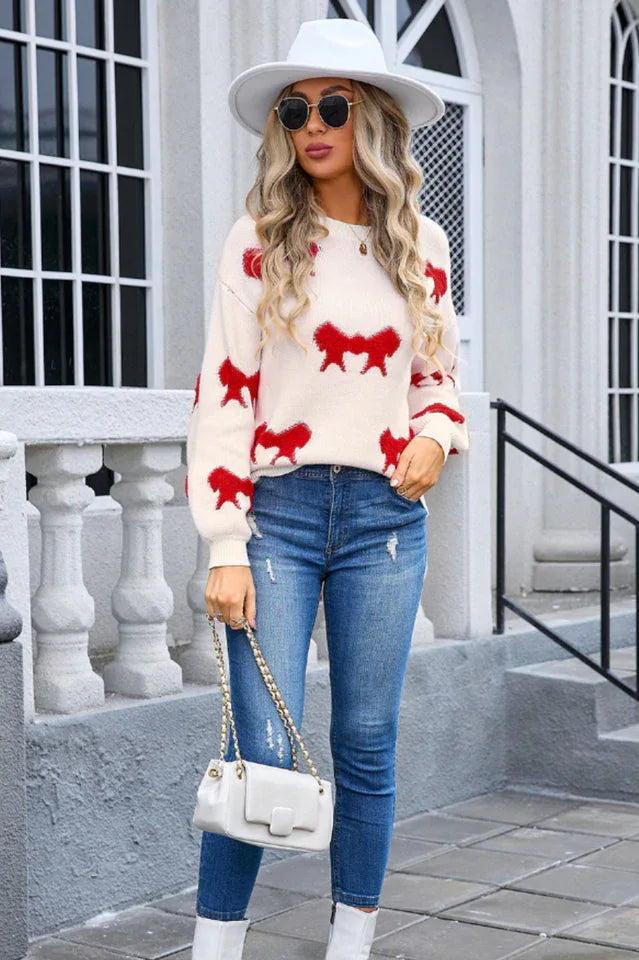 Woman wearing a cream knit sweater with a red bow pattern, blue jeans, and a white handbag