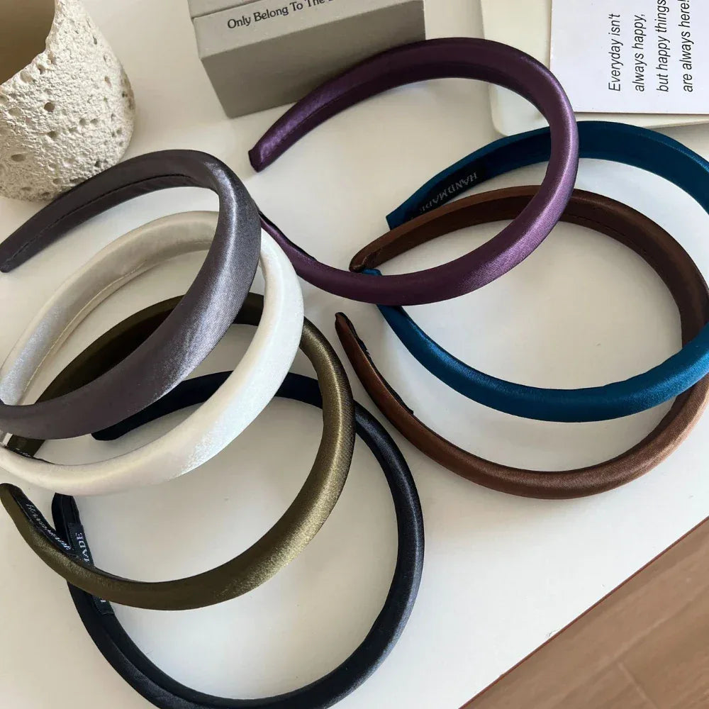 Several padded satin headbands in various colors, including purple, grey, olive green, black, white, teal, and brown, scattered on a white surface