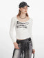 Front view of a woman wearing a white long-sleeved graphic t-shirt, grey distressed jeans with a black fringed belt, and a black beaded necklace.