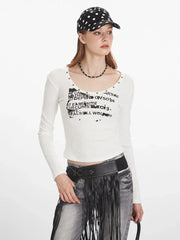 Front view of a woman wearing a white long-sleeved graphic t-shirt, grey distressed jeans with a black fringed belt, and a black beaded necklace.