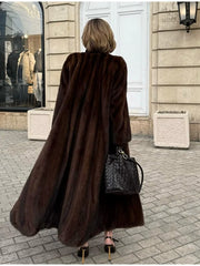 Back view of a woman wearing a long, brown mink fur coat, black leggings, black woven handbag, and brown pointed-toe heels, walking on a cobblestone street.