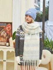 Woman wearing a grey knit sweater with blue and white geometric pattern, a light blue beanie, and a chunky striped scarf, looking forward.