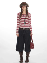 Full body shot of a woman wearing a red and white striped long-sleeved shirt, dark wash wide-leg cropped jeans, a brown belt, brown boots, a dark brown hat with fur trim, and a red handbag.