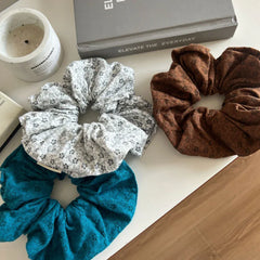 three scrunchies in teal, white with floral pattern, and brown on a white surface with decor