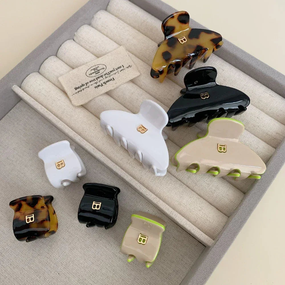 Assorted claw hair clips, including tortoiseshell, black, white, and beige with gold-tone 'B' logos, organized in a grey jewelry tray.