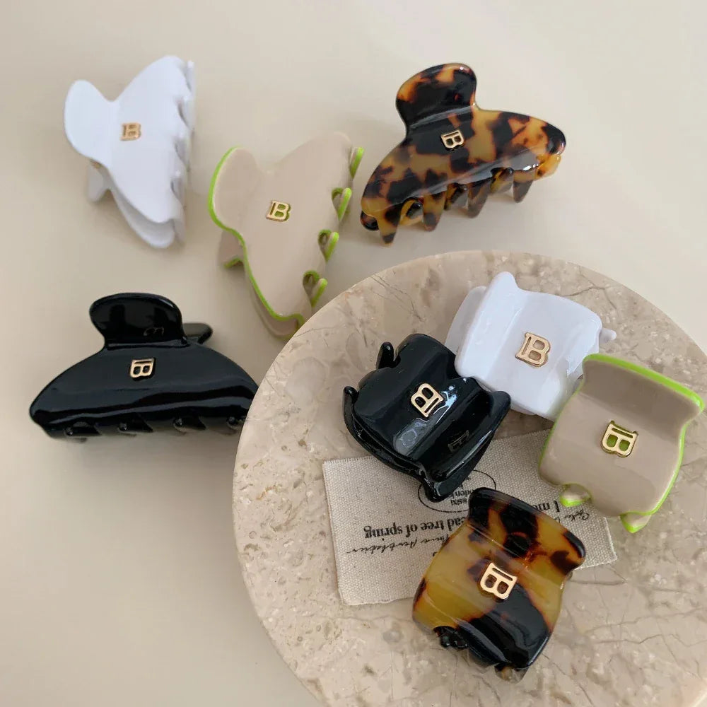 A selection of claw hair clips in different colors and styles, including tortoiseshell, white, black, and beige, arranged on a marble-like surface with fabric.