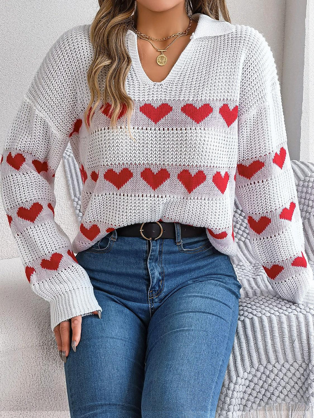 Woman sitting while wearing a white knit sweater with red heart pattern and blue jeans