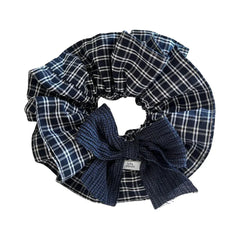 A close-up, overhead shot of a large scrunchie with a navy and white plaid pattern and a dark blue textured bow. A small white tag with black text is visible on the bow.