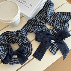 Two scrunchies with navy and white plaid patterns and dark blue textured bows are on a white table next to a white coffee cup and a book.
