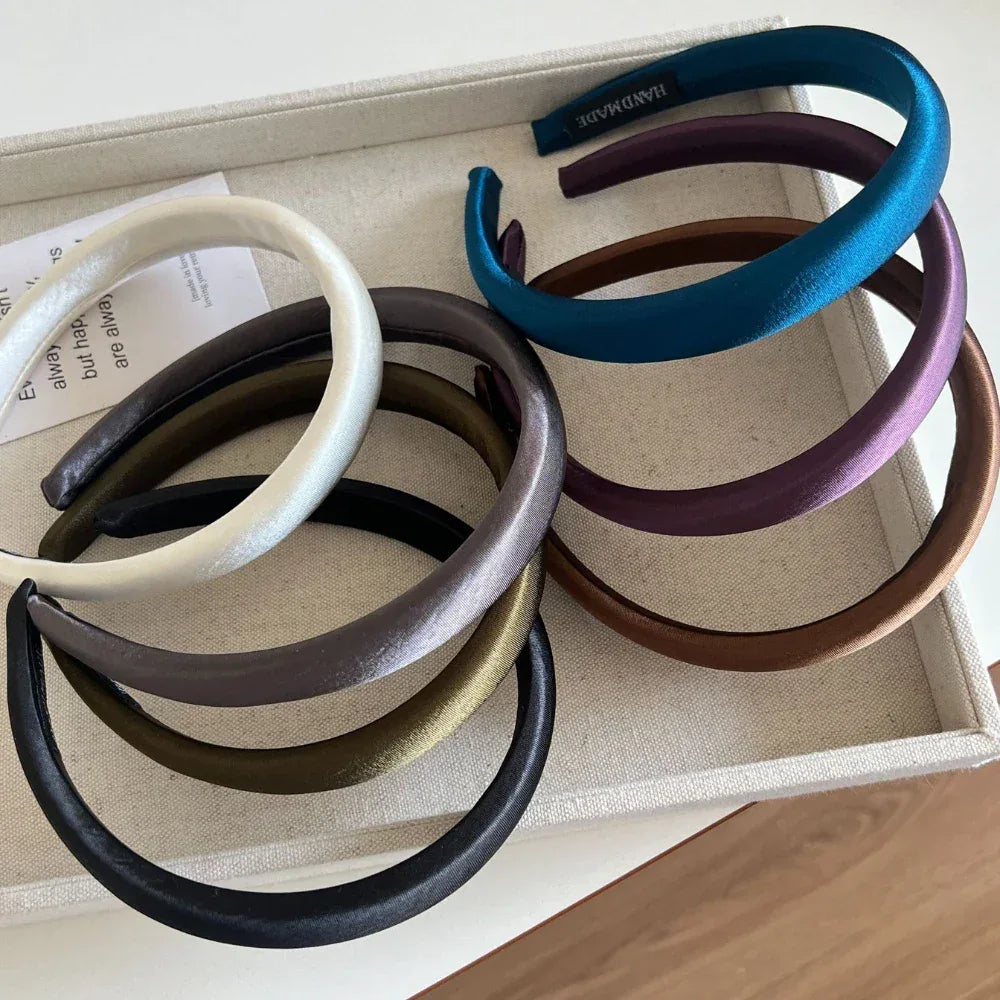 Assorted padded satin headbands in a tray, showing multiple colors including white, grey, olive green, black, purple, teal, and brown