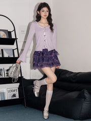Woman posing in a lavender ribbed knit cardigan with lace trim and bow details over a white camisole, paired with a purple tiered ruffled skirt, white knee-high socks, and silver mary jane shoes.