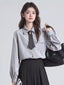Woman wearing a grey pinstripe collared shirt with a dark tie and brooch, a black pleated skirt, and holding a black handbag.