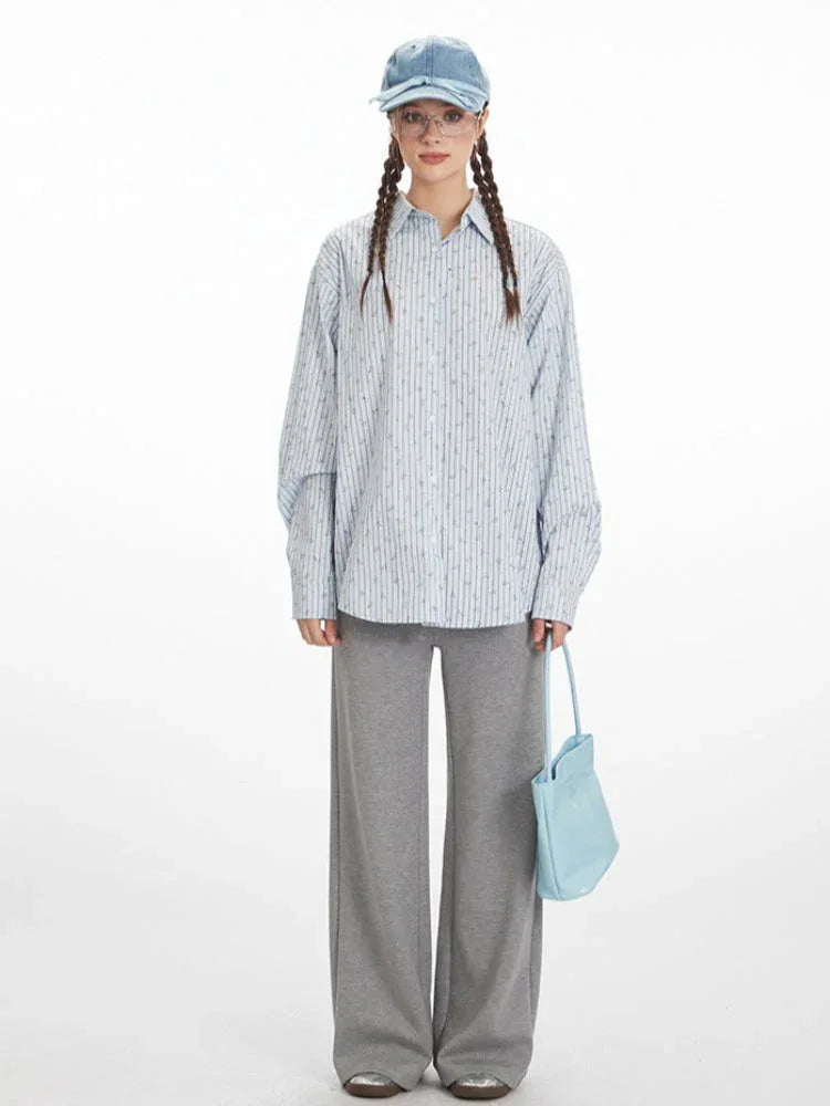 Front view of a woman wearing a light blue pinstripe button-down shirt with a floral print, grey wide-leg pants, and a light blue handbag.