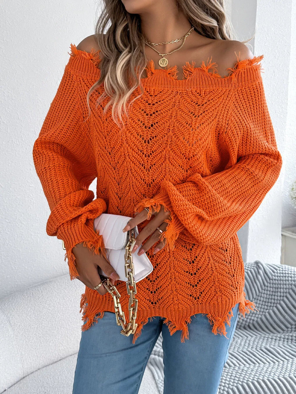 Woman holding a white clutch while wearing an orange distressed off-the-shoulder knit sweater and blue jeans.