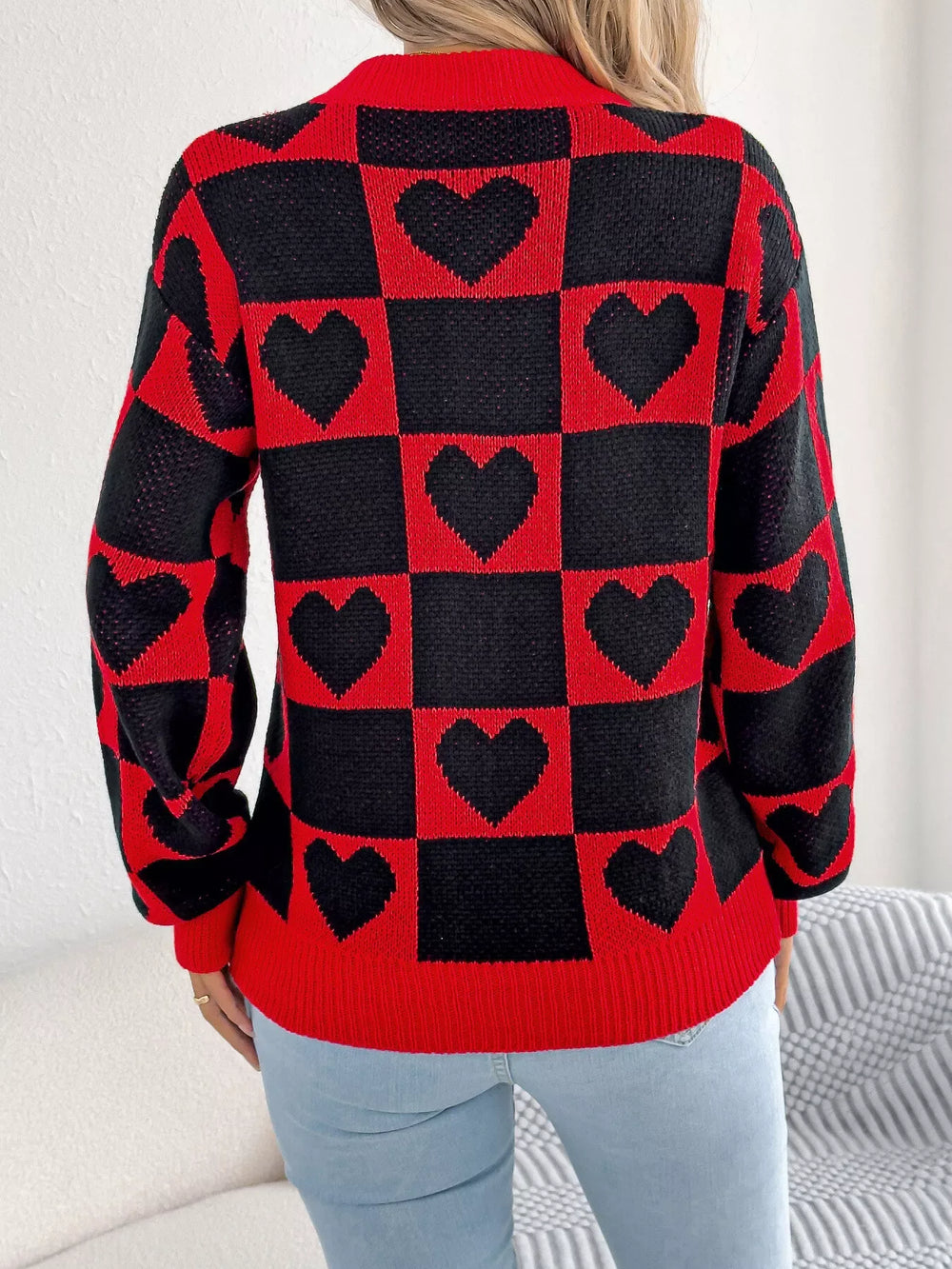 Back view of a woman wearing a red and black checkerboard heart print knitted sweater with light blue jeans