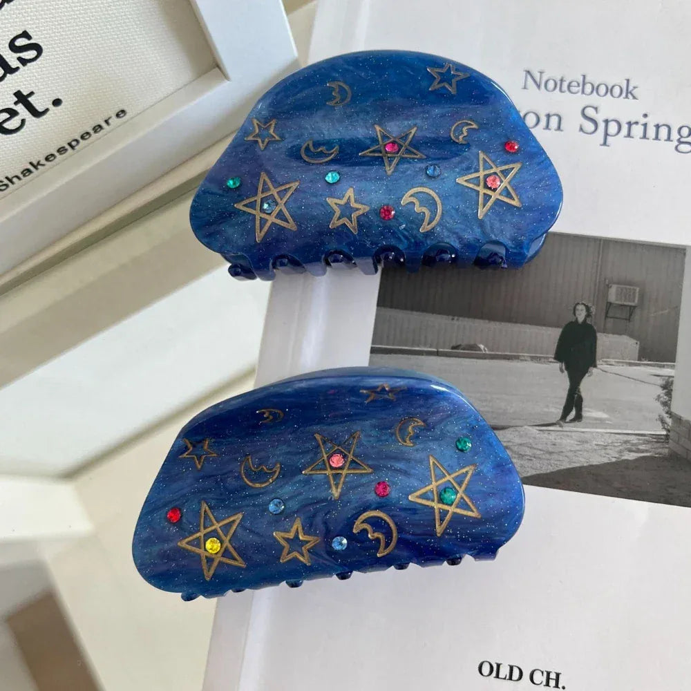 Two blue claw hair clips with celestial patterns are displayed on top of books.
