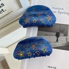 Two blue claw hair clips with celestial patterns are displayed on top of books.