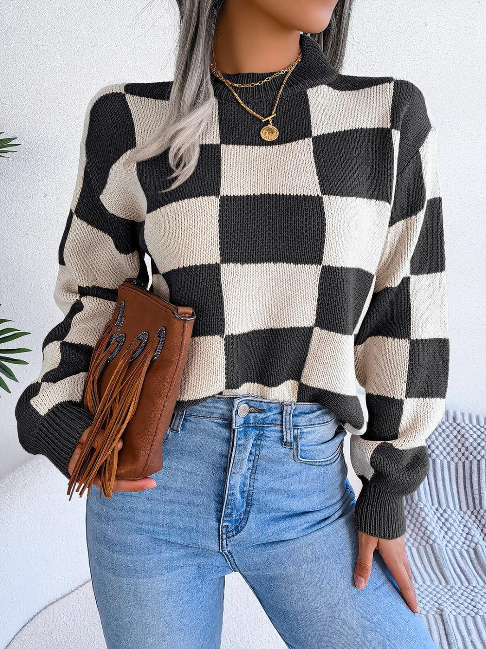 Woman wearing a black and white checkerboard knit sweater with light wash jeans and a brown fringe clutch, front view.