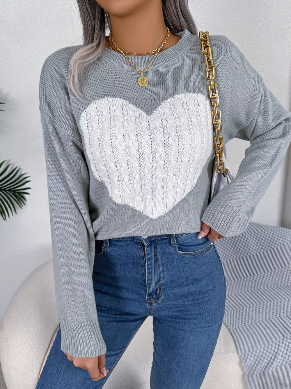 Woman wearing a grey knit sweater with a white cable knit heart graphic, blue jeans, and gold necklaces, holding a white handbag with a gold chain strap.