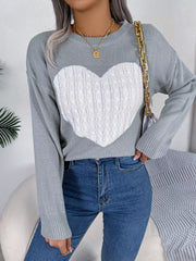 Woman wearing a grey knit sweater with a white cable knit heart graphic, blue jeans, and gold necklaces, holding a white handbag with a gold chain strap.