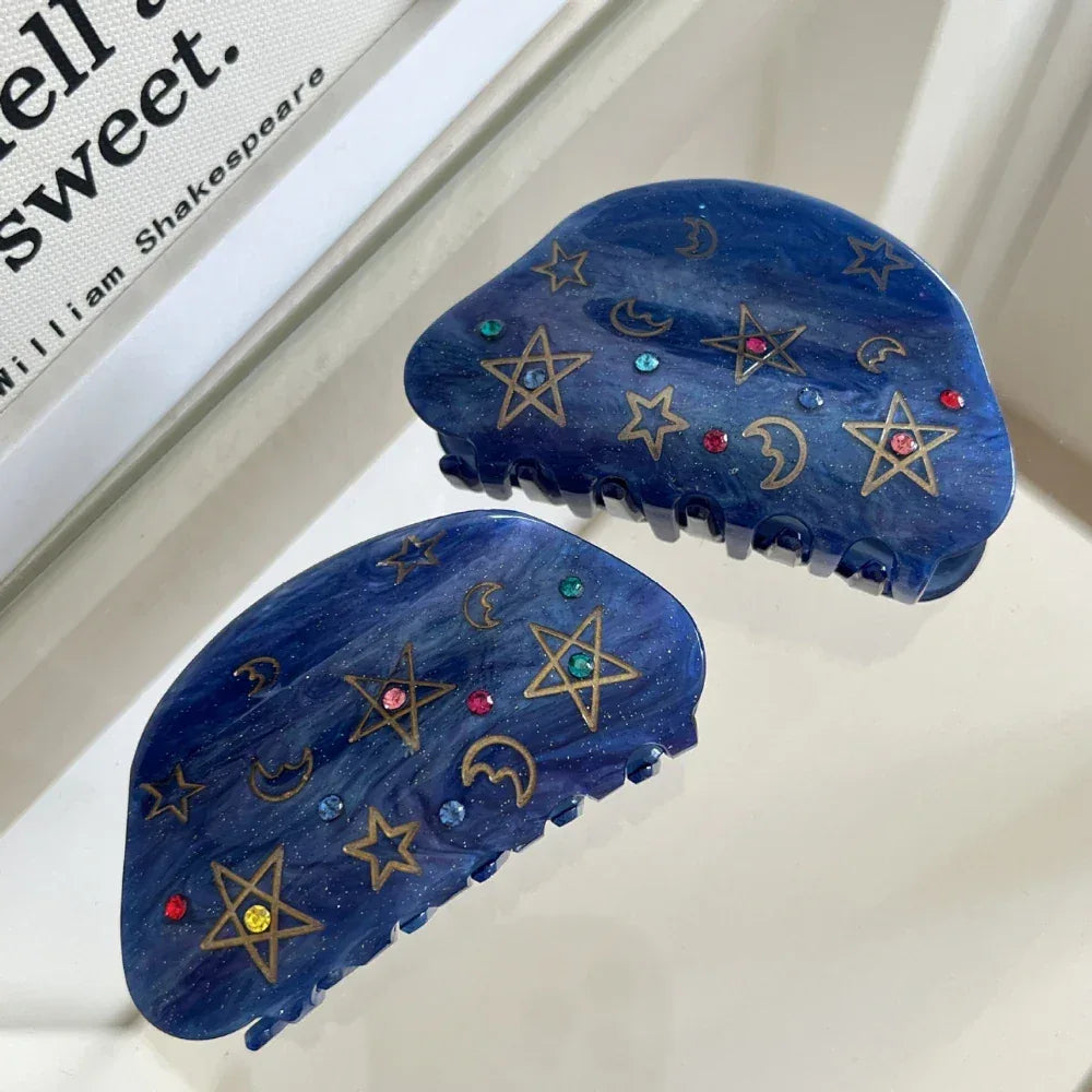 A pair of blue claw hair clips with celestial designs are shown against a reflective surface.