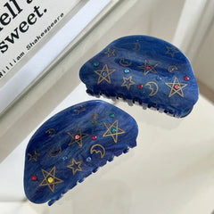 A pair of blue claw hair clips with celestial designs are shown against a reflective surface.