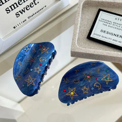 Two blue claw hair clips featuring stars, moons, and gemstones are placed on a surface with books and a tray.