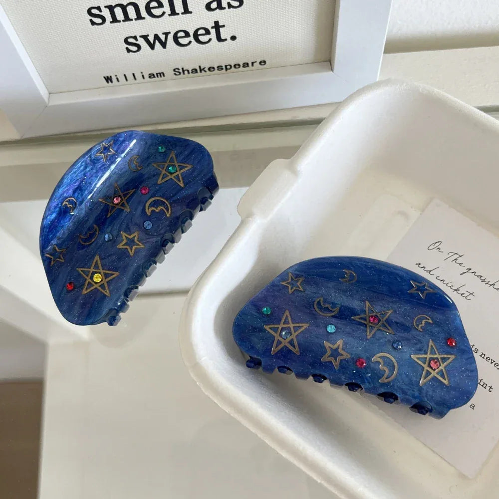 Two blue claw hair clips with celestial motifs are arranged in a white tray.