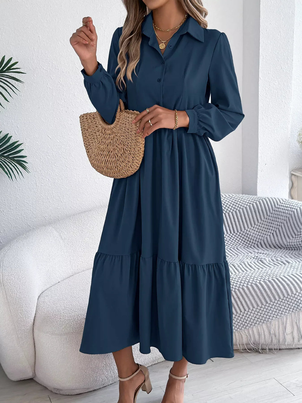 Woman standing holding a straw bag and wearing a long-sleeved blue midi dress with a collared neckline and tiered hem
