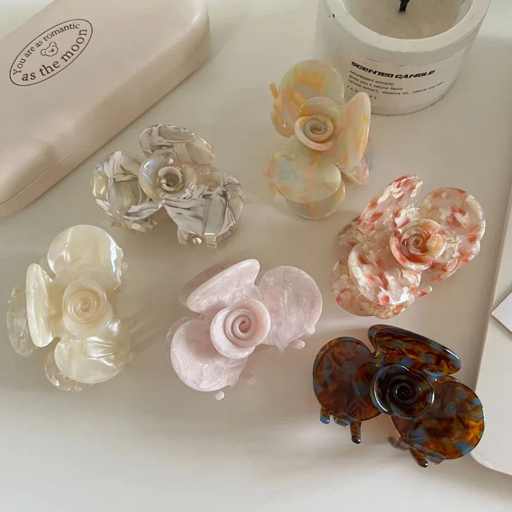 A flat lay of six floral-shaped acetate hair claw clips in different pastel and marbled designs, arranged on a white surface with a candle and box.